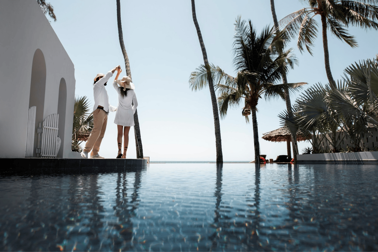 Pareja disfrutando de sus vacaciones bailando junto a la piscina en un apartamento blanco con palmeras.